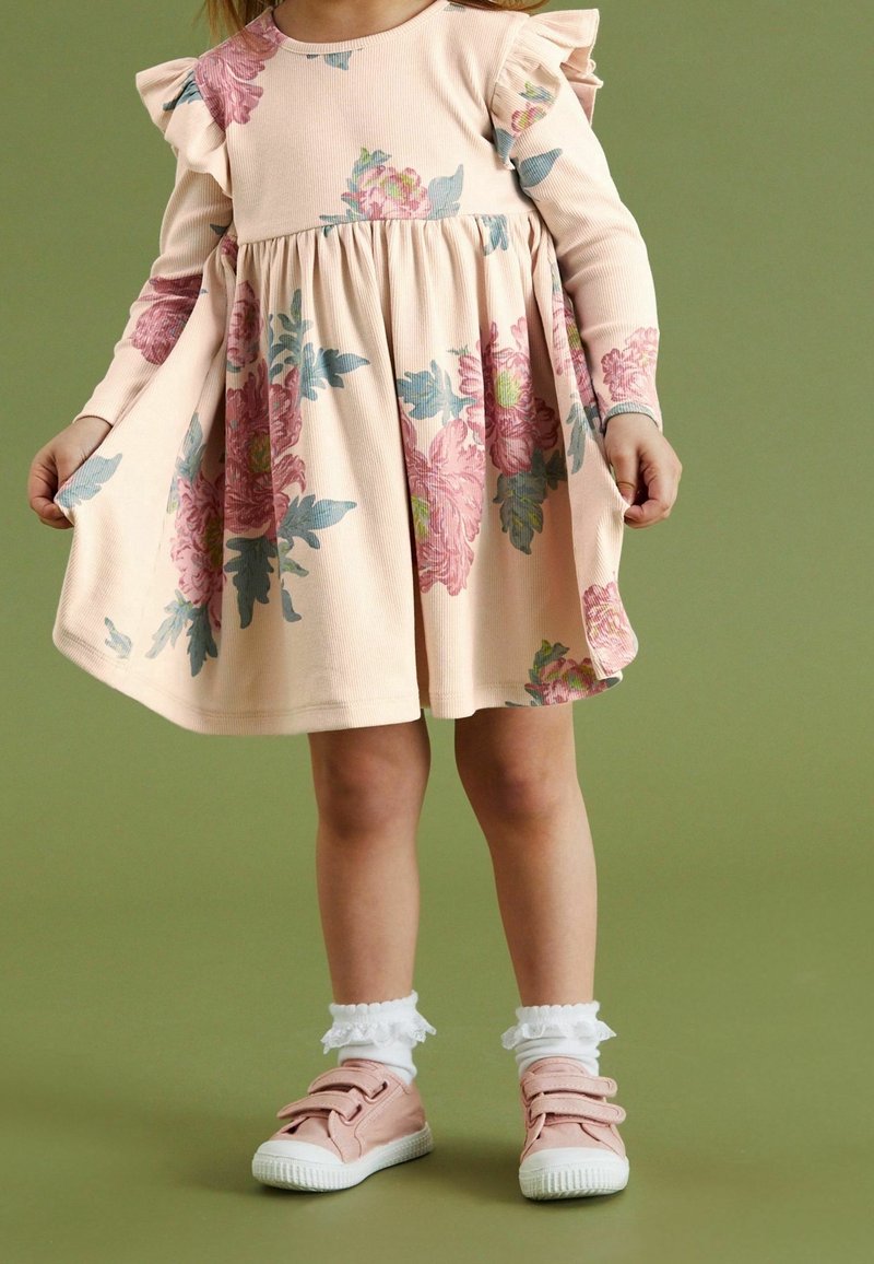 Jeune enfant portant une robe rose à fleurs avec des épaules volantées, des chaussettes blanches à volants et des baskets roses à scratch, debout devant un fond vert.