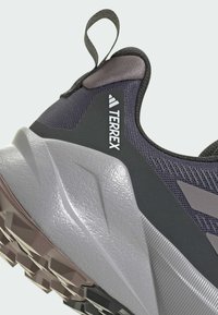 Chaussure de sport grise avec un dessus en maille texturée, un col rembourré et une semelle extérieure en caoutchouc. Accents gris et violet notables et logo Terrex.