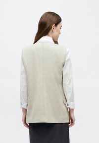 Veste grise clair conçue avec une coupe ajustée, présentant un tissu texturé. Portée sur une chemise blanche aux manches retroussées.