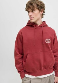 Un jeune homme portant un sweat à capuche rouge avec une poche avant et un petit logo blanc, vêtu de pantalons beiges, regardant sur le côté devant un fond uni.