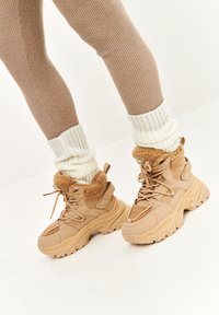 Tenisi bej, cu talpă groasă, guler din blană sintetică, material texturat și talpă din cauciuc. Asortați cu colanți maro cu nervuri și șosete albe din tricot.