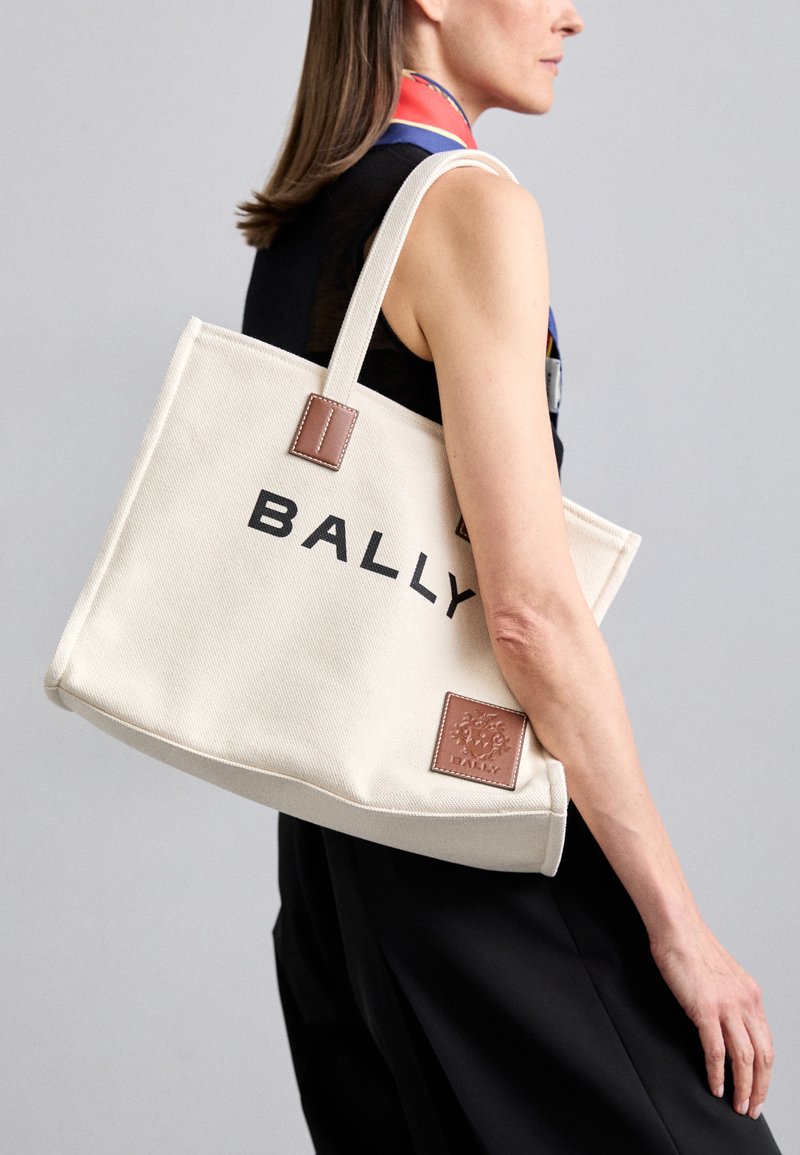 Beige canvas tote bag featuring black "BALLY" logo, brown leather accents, rectangular shape, and sturdy handles. Textured surface.