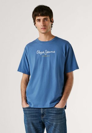 Młody mężczyzna z krótkimi ciemnymi włosami, ubrany w niebieski t-shirt "Pepe Jeans London" i niebieskie dżinsy, z rękami w kieszeniach, na neutralnym tle.