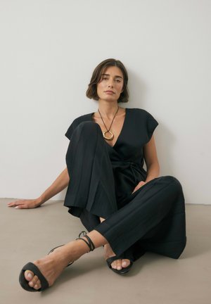 Schwarzer Jumpsuit mit Wickeldesign, kurzen Ärmeln und weitem Bein. Kombiniert mit schwarzen Sandalen mit Riemchen und einer goldenen kreisförmigen Halskette.