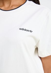 Hvit bomull t-skjorte med rund svart hals. Har svart "adidas"-logo og kløver på venstre bryst. Korte ermer, glatt tekstur.