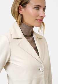 Crème leren blazer met een reverskraag, een enkele witte knoopsluiting en een gladde textuur. Gecombineerd met een aansluitende bruine coltrui.
