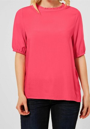 Vrouw die een felroze blouse met korte mouwen en ruches langs de hals draagt en donkerblauwe jeans, staand tegen een effen achtergrond.