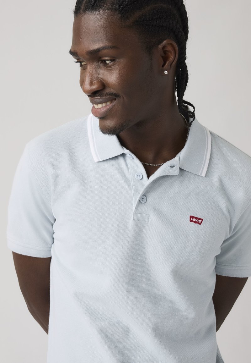 Hombre con el pelo trenzado, lleva una camisa polo azul claro de Levi's, un collar de cadena plateada y un único pendiente de botón, mirando de lado y sonriendo.