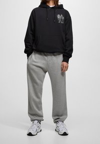 Zwarte hoodie met trekkoorden, voorzak en witte grafische ontwerpen; gecombineerd met grijze joggingsbroek. Model draagt witte sportschoenen.