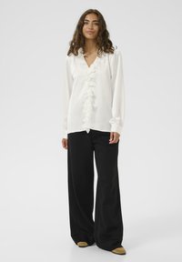 Biała bluzka z falbanami z przodu, długim rękawem i dekoltem w kształcie litery V, w zestawieniu z luźnymi czarnymi spodniami i jasnymi balerinami.
