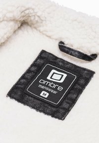 Tessuto bianco soffice con un'etichetta nera che presenta "ombre menswear" e la taglia "M" in testo bianco, cucita saldamente nel capo.