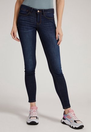 Kobieta ubrana w ciemnoniebieskie obcisłe dżinsy oraz wielokolorowe białe, różowe i fioletowe trampki, stojąca na jednolitym tle.