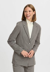 Beige ja mustrihkudus jakker; omadused hõlmavad nurgeliselt lõigatud kraed, ühte nuppu ja tekstuuri, mis tagab vormitud sobivuse.