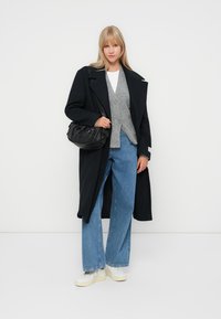 Abrigo largo negro, cárdigan gris, camisa blanca, jeans de pierna ancha azules y zapatillas blancas. Bolso negro con textura arrugada. Fondo claro.