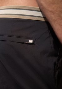 Svarta badbyxor med en randig beige midjeresår och en liten zipficka med en fyrkantig metallaccent. Tyget har en mjuk och len yta.