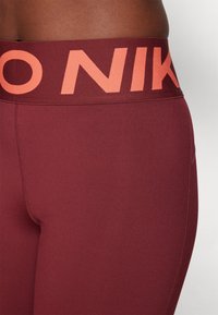 Bordo sportovní legíny s širokým pasem a nápisem "NIKE" v korálové barvě. Hladká textura, přiléhavý st�řih a kontrastní švy.