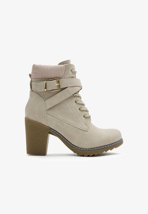 Botín beige con superficie texturizada, parte delantera con cordones, herrajes dorados y un cuello tipo calcetín acanalado. Presenta un tacón grueso y una suela con relieve.