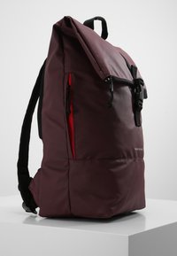 Sac à dos bordeaux avec une surface texturée, doté de sangles noires, d'un rabat supérieur et d'une fermeture éclair rouge sur le côté. Comprend une fermeture à boucle noire.
