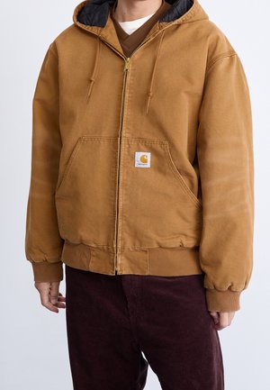 Homme portant une veste à capuche zippée Carhartt marron sur un t-shirt marron à col en V et un pantalon en velours côtelé marron foncé.