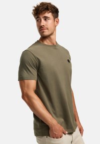 Olivegrünes Baumwoll-T-Shirt mit rundem Halsausschnitt, kurzen Ärmeln und einem kleinen schwarzen Patch-Detail auf der Brust. Glatte Textur.