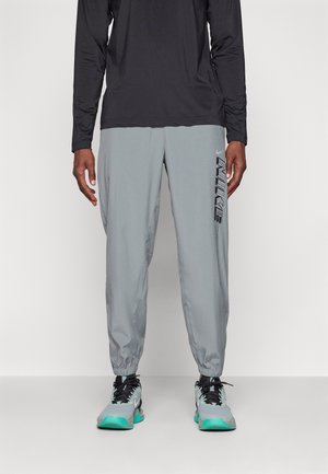 Mann trägt graue Nike-Jogginghosen mit schwarzem Logo auf dem rechten Bein, langärmliges schwarzes Oberteil und graue Sneakers mit türkisfarbenen Akzenten.