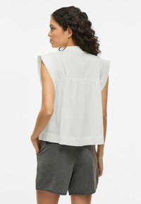 Witte mouwloze top met geplooide achterkant, geplooide schouders en een losse pasvorm, gecombineerd met donkerdenim shorts. Gladde textuur en eenvoudig ontwerp.