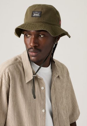 Olijfgroene emmerhoed met een zwart gestikt label, verstelbare kinband en een gestructureerde afwerking, gedragen bij een geruit overhemd.