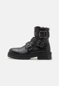 Bota de cuero negra hasta el tobillo con tres correas ajustables y hebillas plateadas. Presenta una punta redondeada y una suela de goma texturizada para mayor tracción.