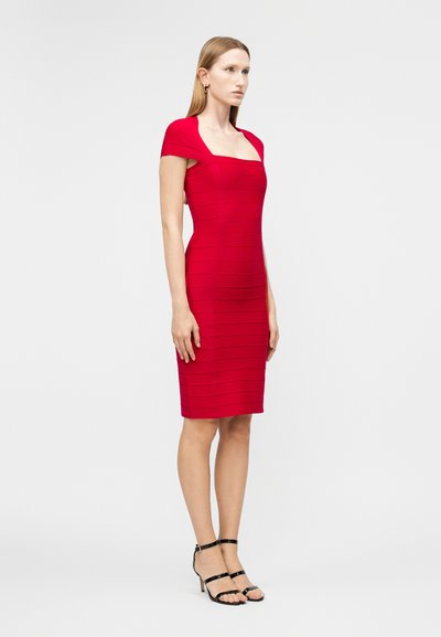 Žena stojící bokem, oblečená do přiléhavých červených šatů délky po kolena s krátkými rukávy a černými sandály na podpatku, na jednotném pozadí.