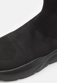 Czarne lekko dzianinowe sneakersy z teksturowanym wierzchem, oddychającymi sekcjami z siateczki oraz grubą gumową podeszwą o zakrzywionym kształcie.