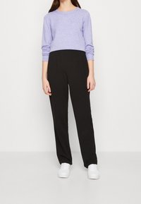 Persona con un suéter de manga larga de color púrpura claro, pantalones negros de cintura alta y rectos, y zapatillas blancas, de pie contra un fondo liso.