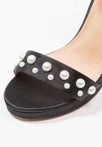 Czarne satynowe sandały na wysokim obcasie z szerokim paskiem ozdobionym różnej wielkości białymi perłami, gładką matową podeszwą i neutralną wyściółką wewnętrzną.