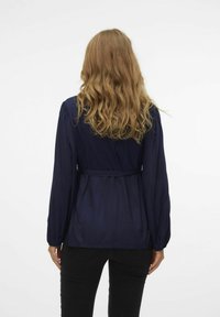 Blusa azul marino con mangas largas, detalles fruncidos en los hombros y cintura ceñida con cinturón. Textura suave, tejido ligero, presenta un estilo peplum.