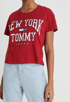 Osoba w czerwonym t-shircie z krótkim rękawem z napisem "New York Tommy Jeans" i jasnoniebieskich dżinsach z wysokim stanem na jednolitym tle.