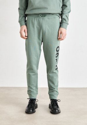 Green cotton sweatpants with black logo print on the left leg, featuring an elastic waistband and slightly tapered legs. Worn with black shoes.