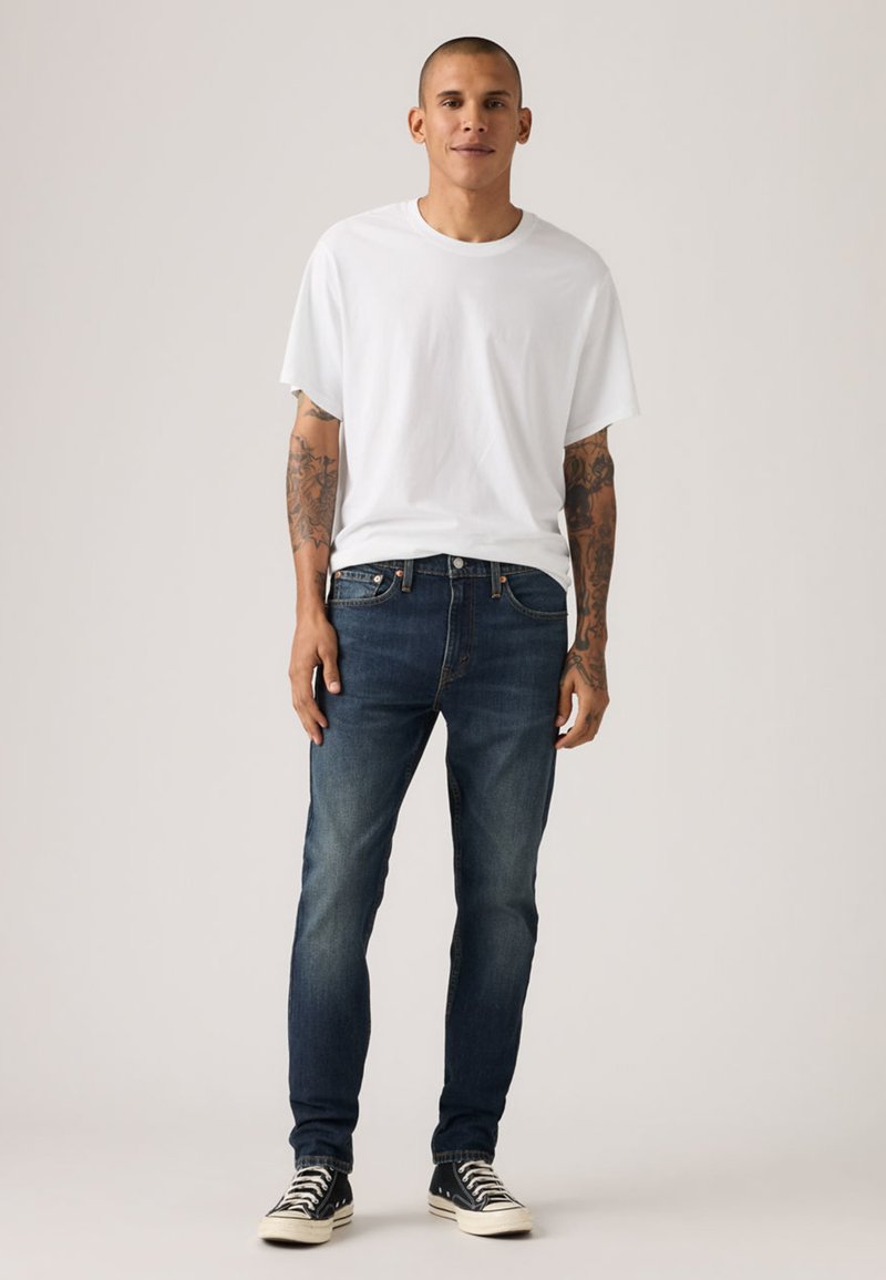 T-shirt en coton blanc, jean denim bleu et baskets noires. Le jean présente une coupe ajustée et un délavage subtil, avec un style classique à cinq poches.
