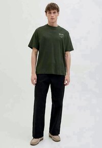 Dark green cotton t-shirt with a crew neckline and short sleeves, featuring minimal white text on the chest, paired with black pants.