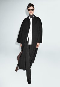 Donna che indossa un lungo cappotto nero sopra una camicia bianca e un dolcevita nero, pantaloni neri, occhiali da sole, che porta una borsa, mentre cammina su uno sfondo semplice.