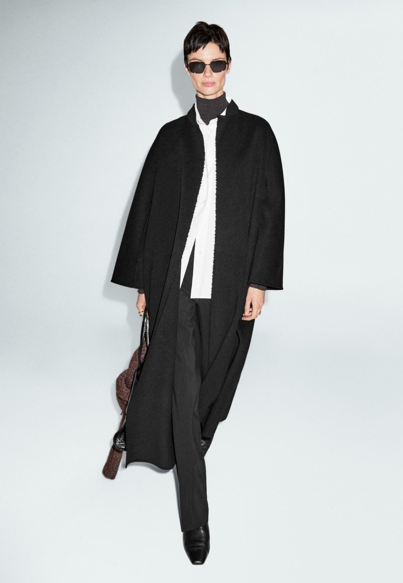 Donna che indossa un lungo cappotto nero sopra una camicia bianca e un dolcevita nero, pantaloni neri, occhiali da sole, che porta una borsa, mentre cammina su uno sfondo semplice.