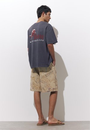 Muž stojí, nosí tmavosivé tričko s červeným textom a béžové krátke nohavice so vzorom, ruky v kapsách, opálené sandále na sivom podlahe.