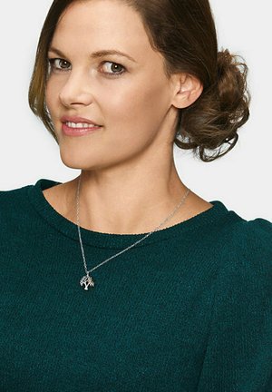 Silberne Halskette mit Baum-Anhänger, die über einem dunkelgrünen strukturierten Pullover getragen wird. Der Pullover hat einen runden Ausschnitt und leicht gepuffte Ärmeln.
