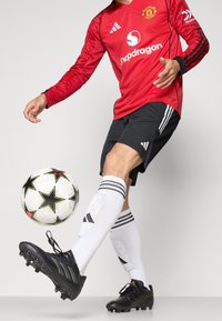 Maglia rossa a maniche lunghe con dettagli neri e logo, pantaloni corti neri, calze bianche a ginocchio con righe e scarpini neri, mentre dribbla un pallone da calcio a fantasia.