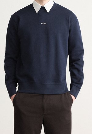 Mann trägt marineblauen Sweatshirt mit weißem „BOSS“-Schriftzug, weißes Hemd mit Kragen darunter und dunkelbraune Hose, Hände in den Taschen, vor einfarbigem Hintergrund.