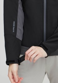 Crna i siva softshell jakna s prednjim zatvaračem i čičak rukavima; sadrži vez "W-PRO 8000" na rukavu. Glatka tekstura.