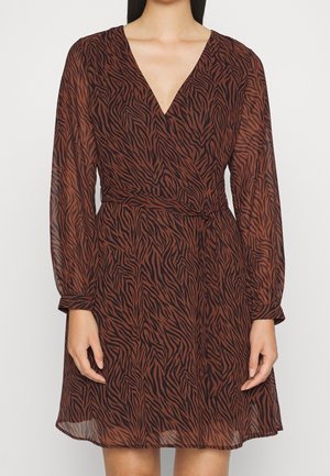 Vrouw die een bruine en zwarte wikkeljurk met zebraprint draagt, met lange mouwen en een V-hals, gestrikt in de taille met een stoffen riem.