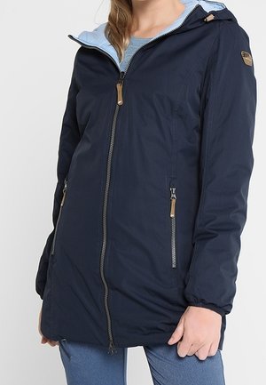 Kobieta ubrana w granatową długą kurtkę z kapturem, z dwoma przednimi kieszeniami na zamek i jasnoniebieskim wewnętrznym podszyciem.