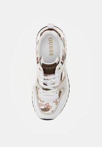 Zapatilla blanca con detalles en patrón marrón, que presenta un diseño de cordones, una suela acolchada y la marca en dorado "GUESS" en la lengüeta.