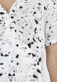 Gros plan sur un tissu blanc avec des taches irrégulières noires, grises et beiges, avec de petits boutons ronds argentés sur un vêtement style portefeuille.