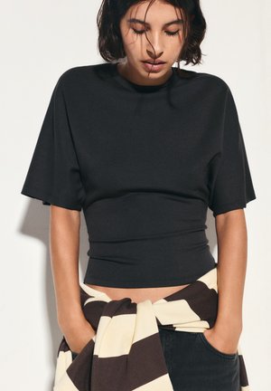 Femme aux cheveux courts et foncés portant un top noir ajusté et un pull rayé noué autour de la taille, regardant vers le bas, les mains dans les poches.