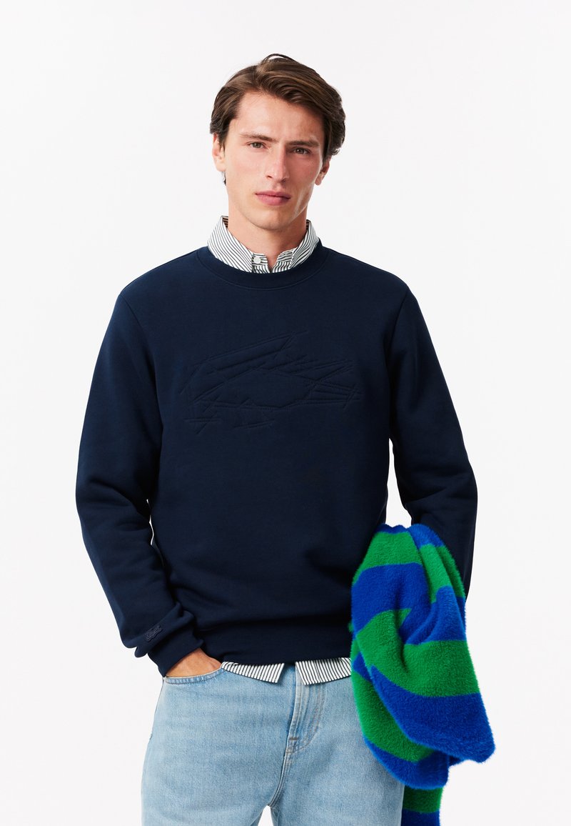 Felpa in cotone blu navy con logo in rilievo, colletto tondo e polsini a coste, abbinata a una coperta in fleece a strisce blu e verde.